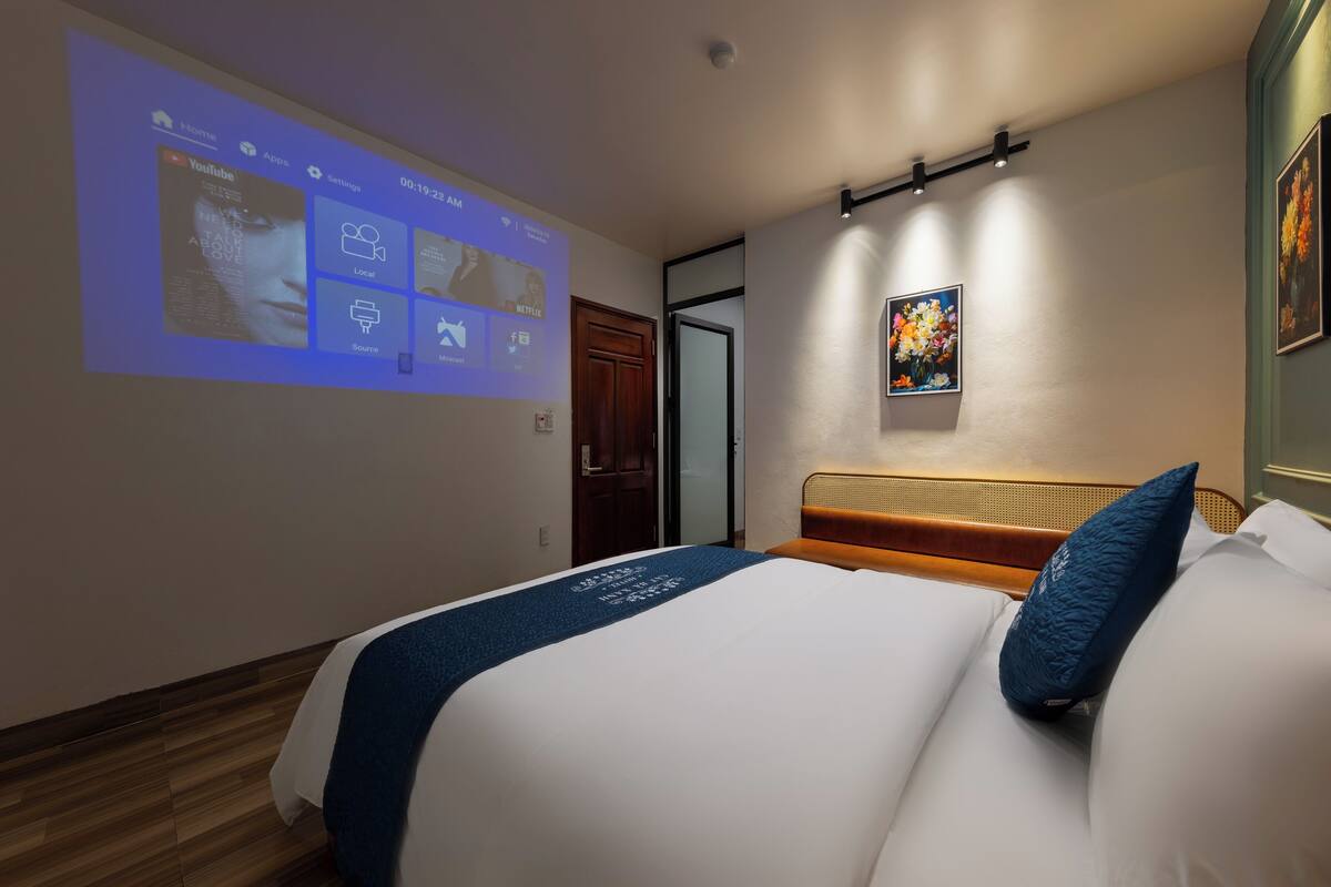 Ein Hotelzimmer mit Bett, einem Projektorbildschirm, der eine YouTube-Oberfläche zeigt, einem Bild an der Wand und einem Holzboden.