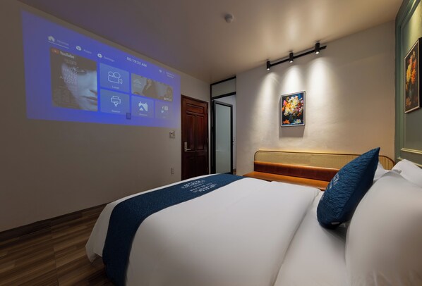 A hotel room with a bed, a projector screen displaying a YouTube interface, a painting on the wall, and a wooden floor. - Cat Ba Xanh Spring Garden Hotel  (Hai Phong)
