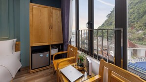 Une chambre comprenant un lit, un bureau avec un ordinateur portable, un petit réfrigérateur et une fenêtre donnant sur des immeubles et des collines.