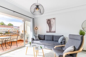 A modern living room with a grey sofa, wooden armchairs, a dining table with chairs, and a large window with a view of buildings.