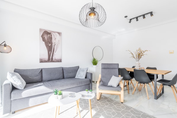 Una sala de estar moderna con un sofá gris, una mesa de comedor con sillas, una lámpara colgante y un espejo montado en la pared.