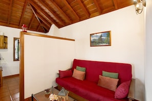 A red sofa with cushions, a wooden coffee table with flowers, a framed picture on the wall, and a wooden ceiling.