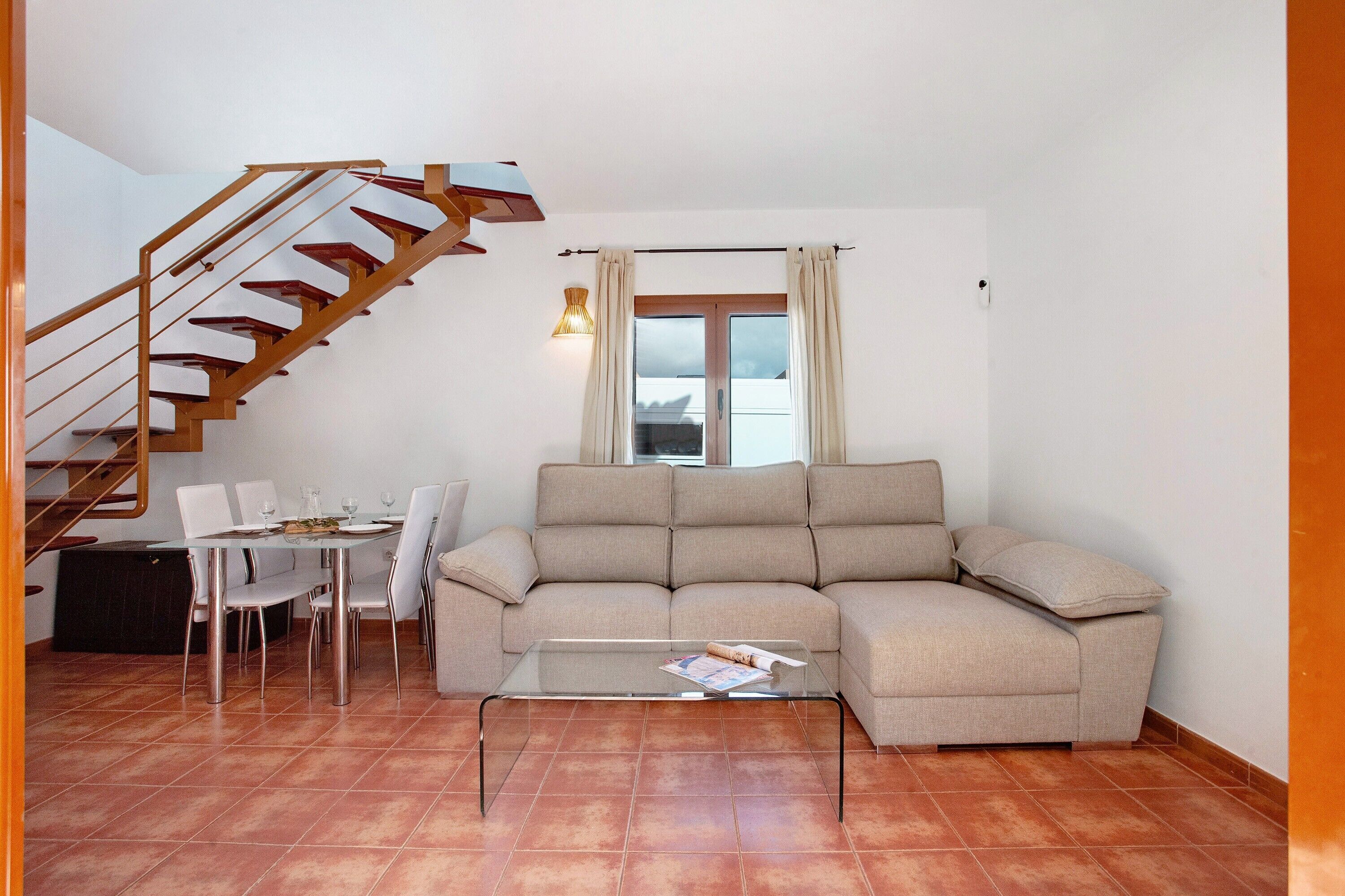 A living room with a sofa, a glass coffee table, a dining table with chairs, and a staircase.