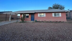 A single-story brick house with a blue door, a covered patio, and a gravel driveway.
