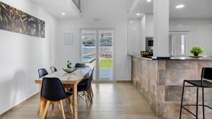 Une cuisine moderne avec un îlot central en pierre, un coin repas doté d’une table en verre, et une vue sur une piscine et un espace verdoyant.