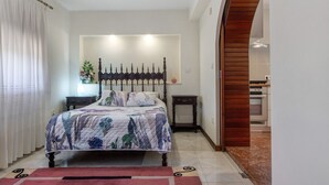 Un dormitorio con cama, mesita de noche, lámpara, planta en maceta y vista a la cocina a través de un arco.