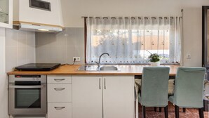 Cocina compacta con horno integrado, microondas, lavamanos y ventana con cortina. Hay dos sillas y una mesita.