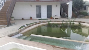En swimmingpool med et lille vandelement, en trappe og en dør der fører til et overdækket område.