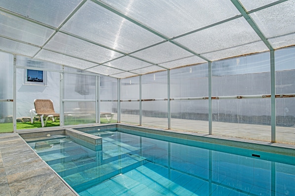 A covered swimming pool with clear glass walls, a bench, and a small window.