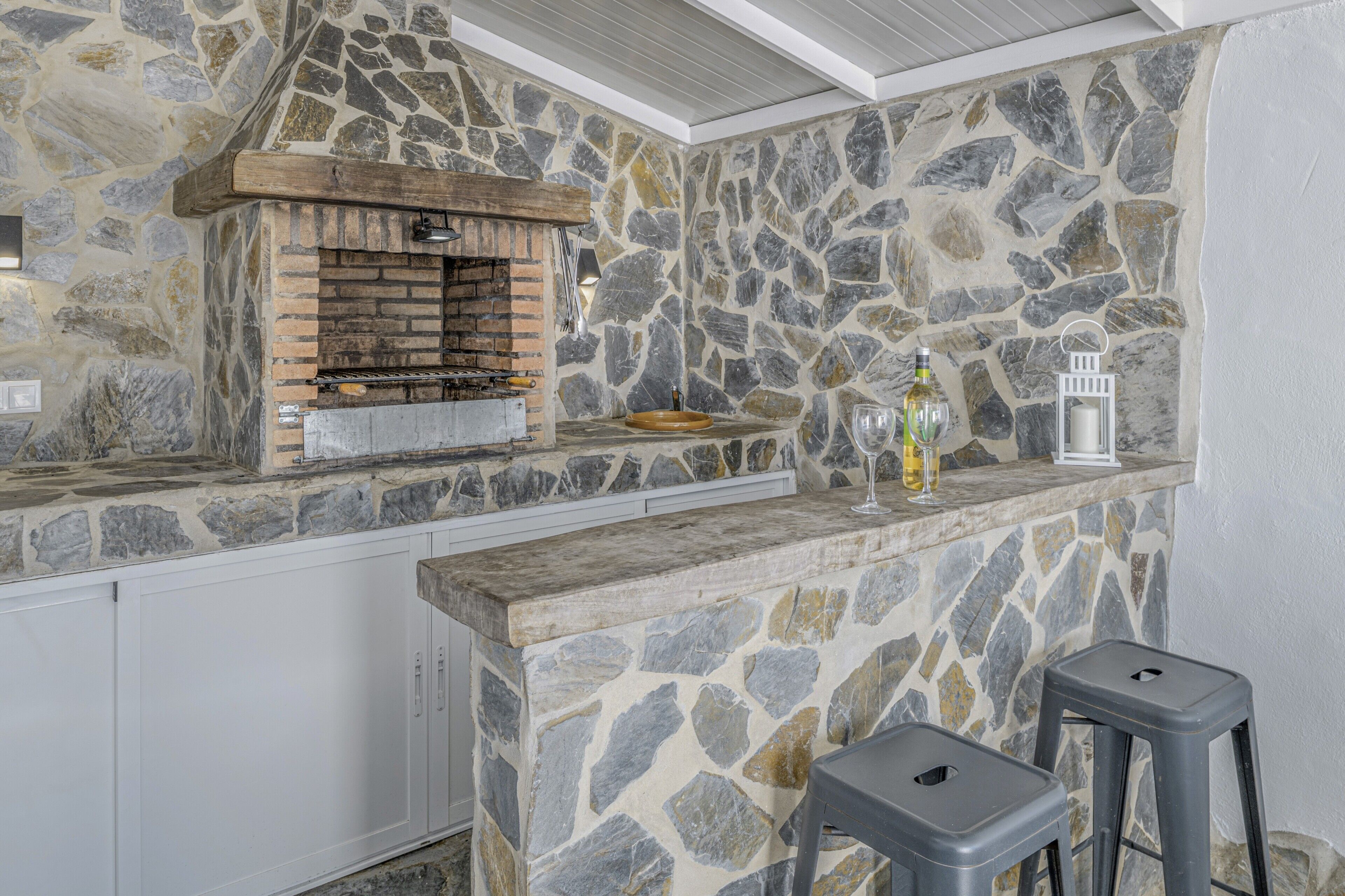 Un mur en pierre agrémenté d’un barbecue intégré et d’un espace bar, comprenant des tabourets et un porte-verres à vin.