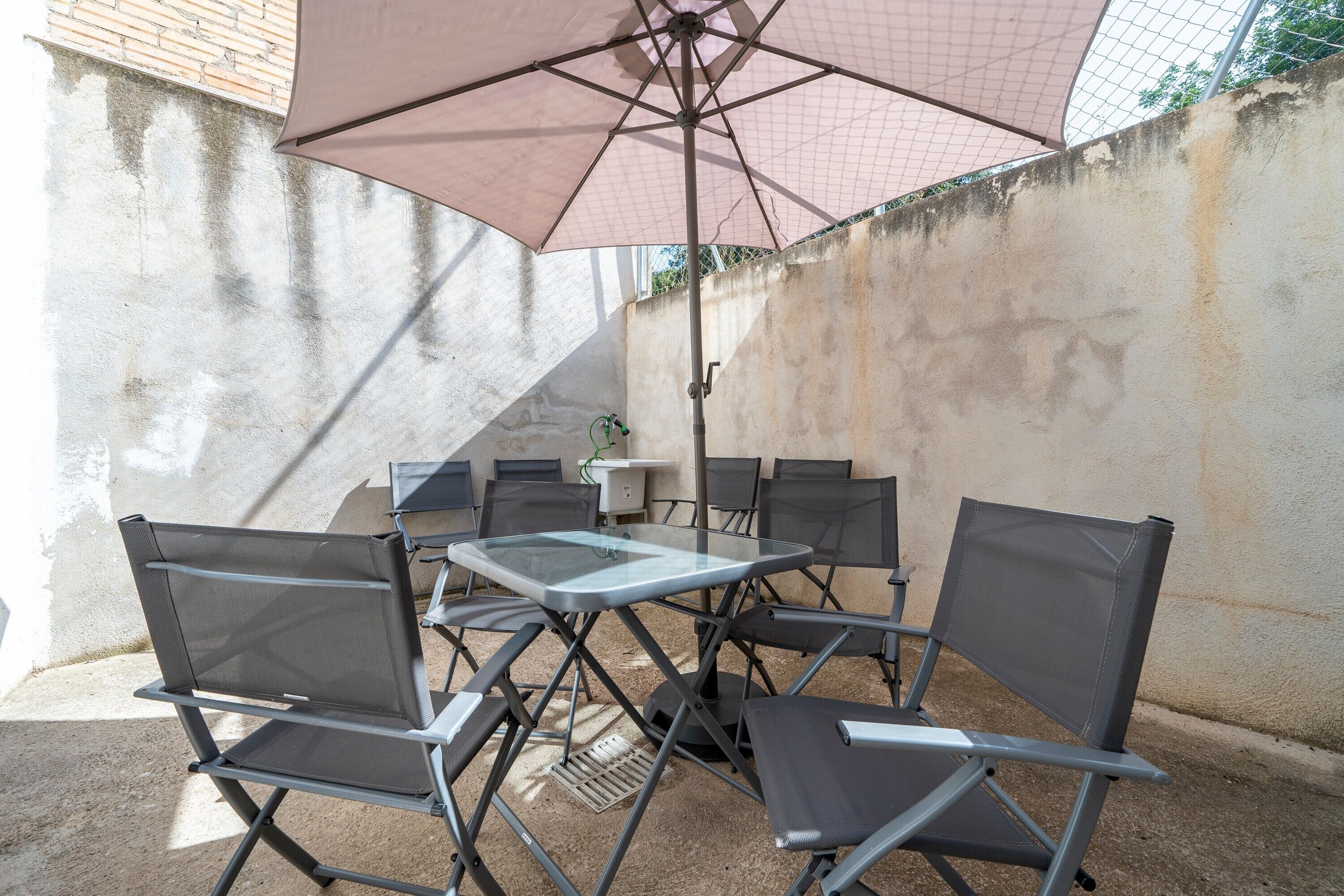 Een terras buiten met een tafel, stoelen, een grote parasol en een betonnen muur.