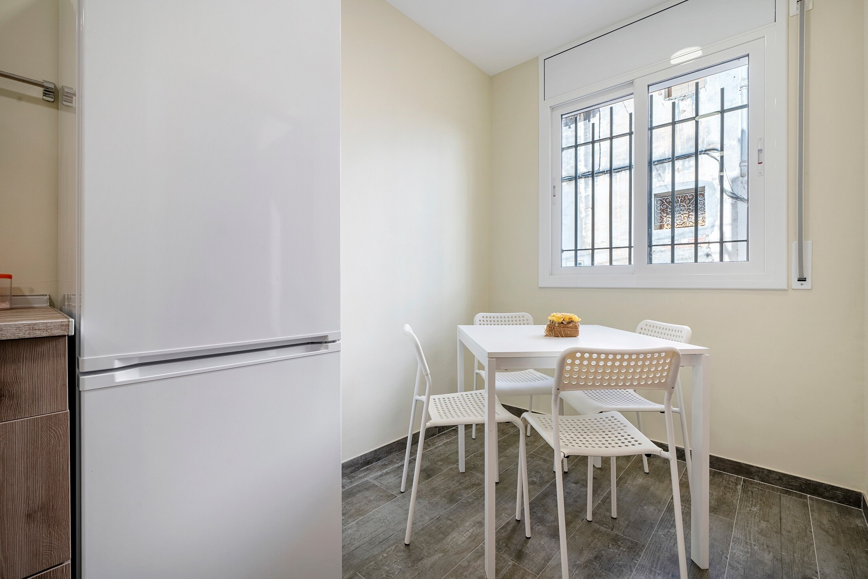 A compact kitchen with a white refrigerator, a dining table with four white chairs, and a window with bars.
