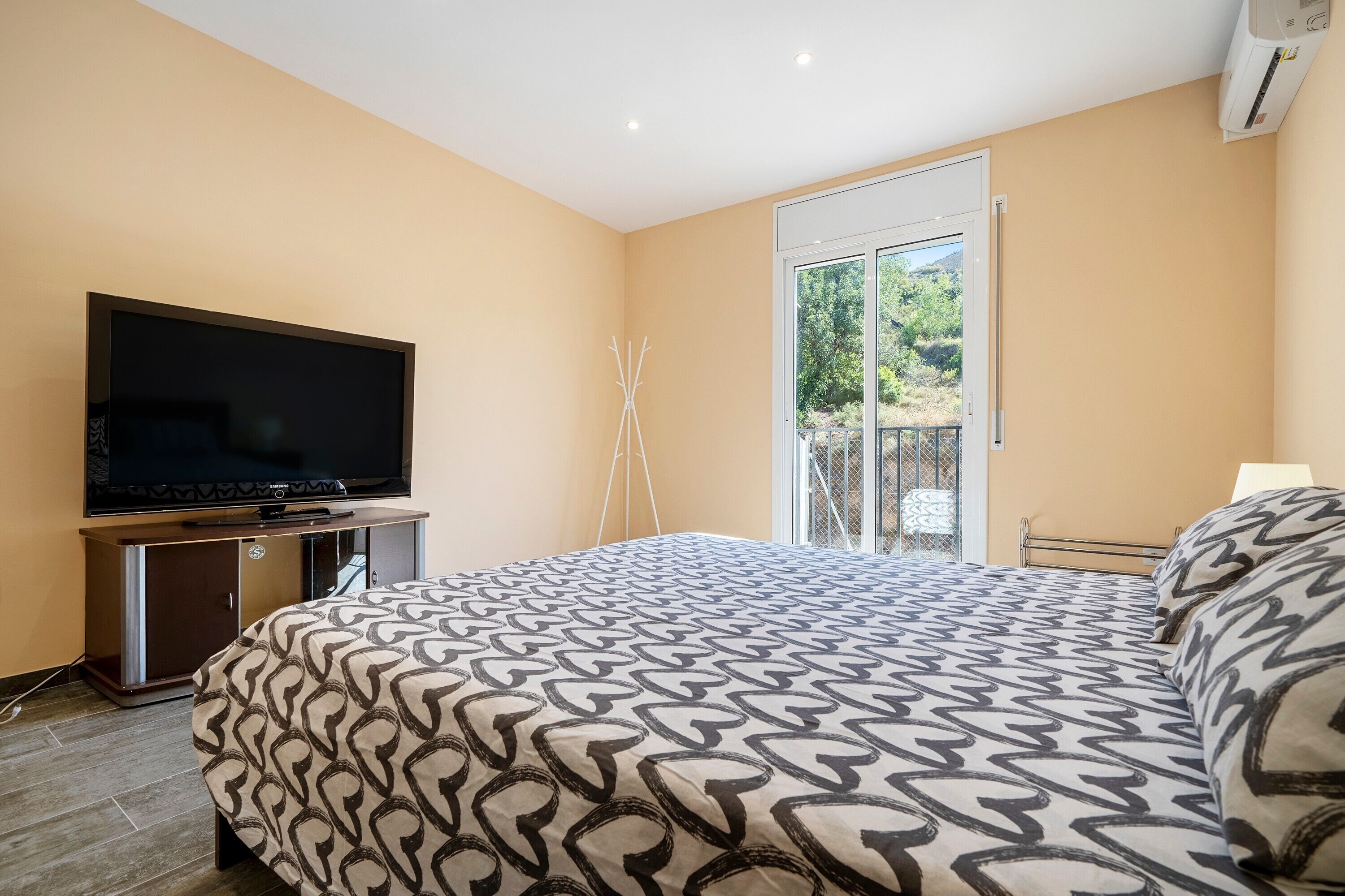 Une chambre à coucher avec un lit, un téléviseur posé sur un meuble, une fenêtre donnant sur un espace verdoyant et un appareil de climatisation.