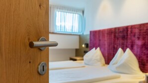 Een houten deur met een sleutelgat en een deurklink, een bed met witte lakens en kussens, een raam met zonwering, en een paars hoofdeinde.