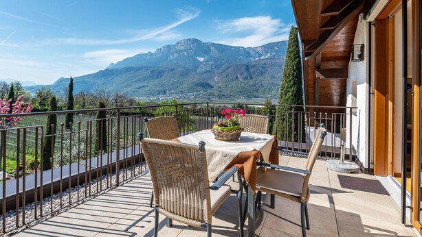 Een balkon met rieten stoelen, een tafel met een bloemenmand en uitzicht op een berg.