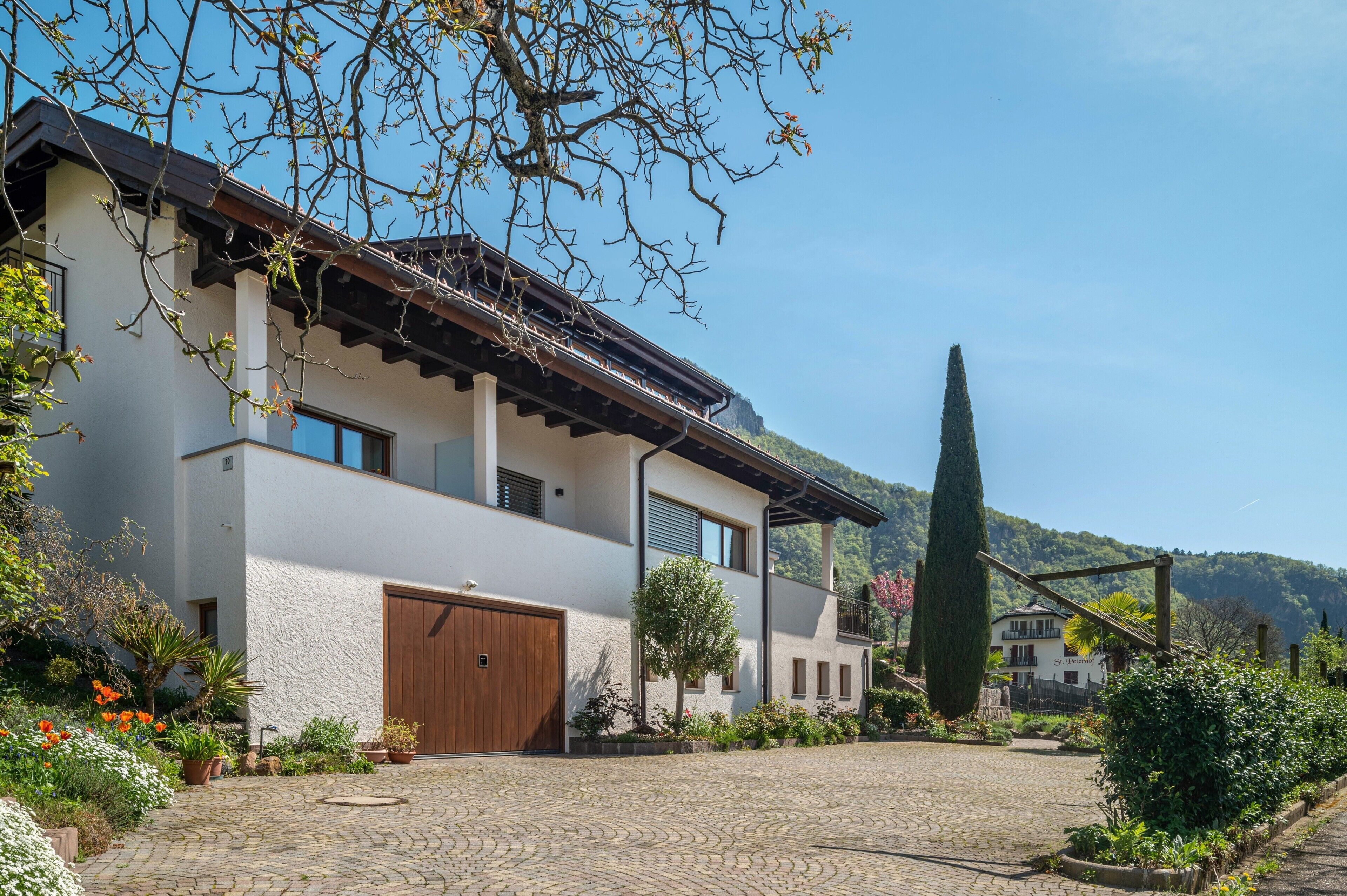 Una casa moderna de dos pisos con un camino pavimentado, una puerta de garaje de madera y un jardín bien cuidado con diversas plantas y flores.