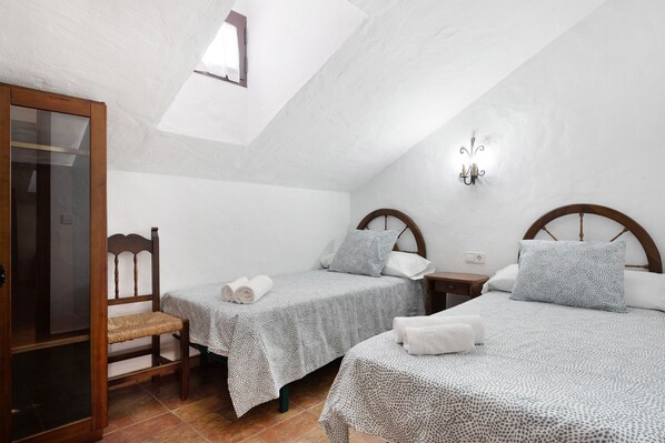 A bedroom with two beds, a wooden chair, a small table, and a wall-mounted light fixture.