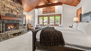 A bedroom with a stone fireplace, a large bed, a window with a view of trees, and a wooden ceiling.
