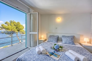A bedroom with a large window, a bed with a patterned bedspread, two pillows, a nightstand with a lamp, and a view of the sea.