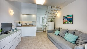 A modern living space with a kitchen, a grey sofa with teal cushions, a flat-screen TV, and a staircase leading to an upper level.