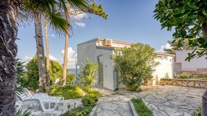 A stone pathway leading to a white building with a tiled roof, surrounded by greenery and palm trees.