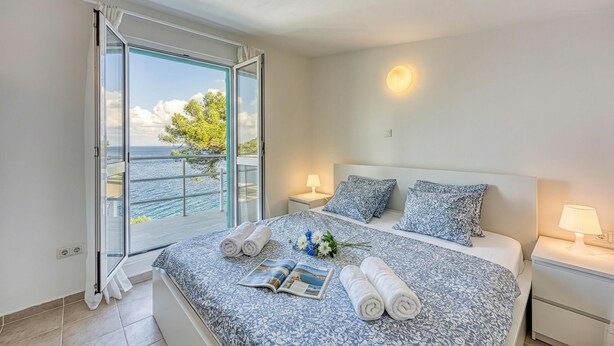 A bedroom with a large window, a bed with a patterned bedspread, two bedside tables with lamps, and a view of the sea.