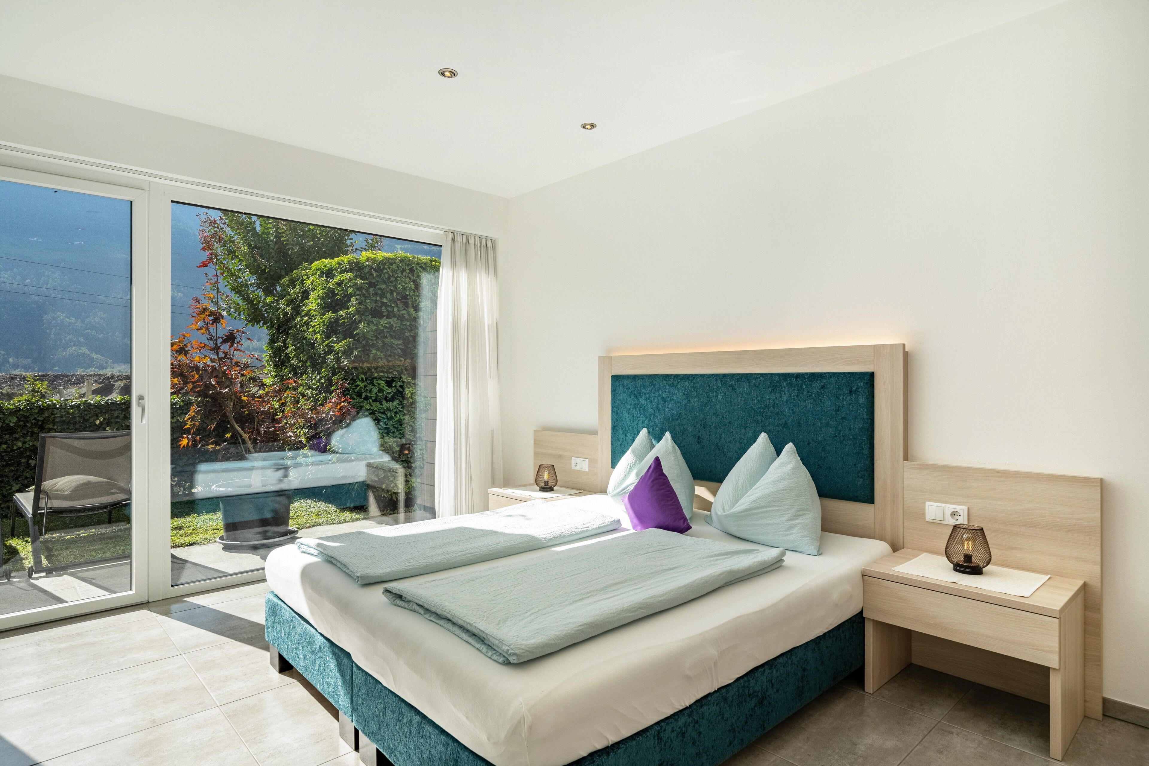 Une chambre avec un lit, des tables de chevet, une vue à travers une porte-fenêtre coulissante et une petite table avec des chaises à l’extérieur.