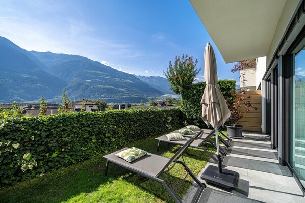 A balcony with a view of mountains, a hedge, and a lounge chair with a cushion.