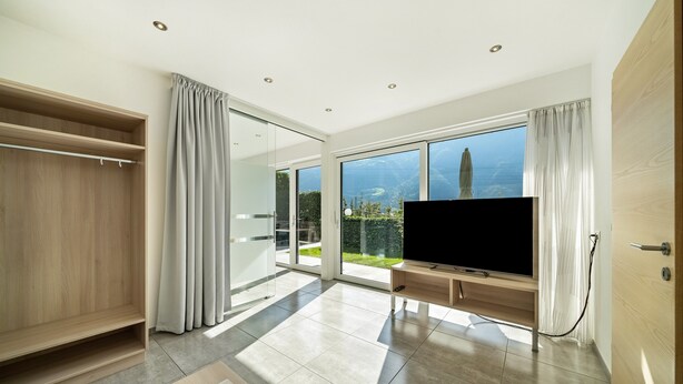 A modern living room with a large TV, sliding glass doors to a balcony, and a wooden wardrobe. - Apartment "Weinreichhof Marie & Max" with Mountain View, Private Terrace & Wi-Fi (Ciardes)