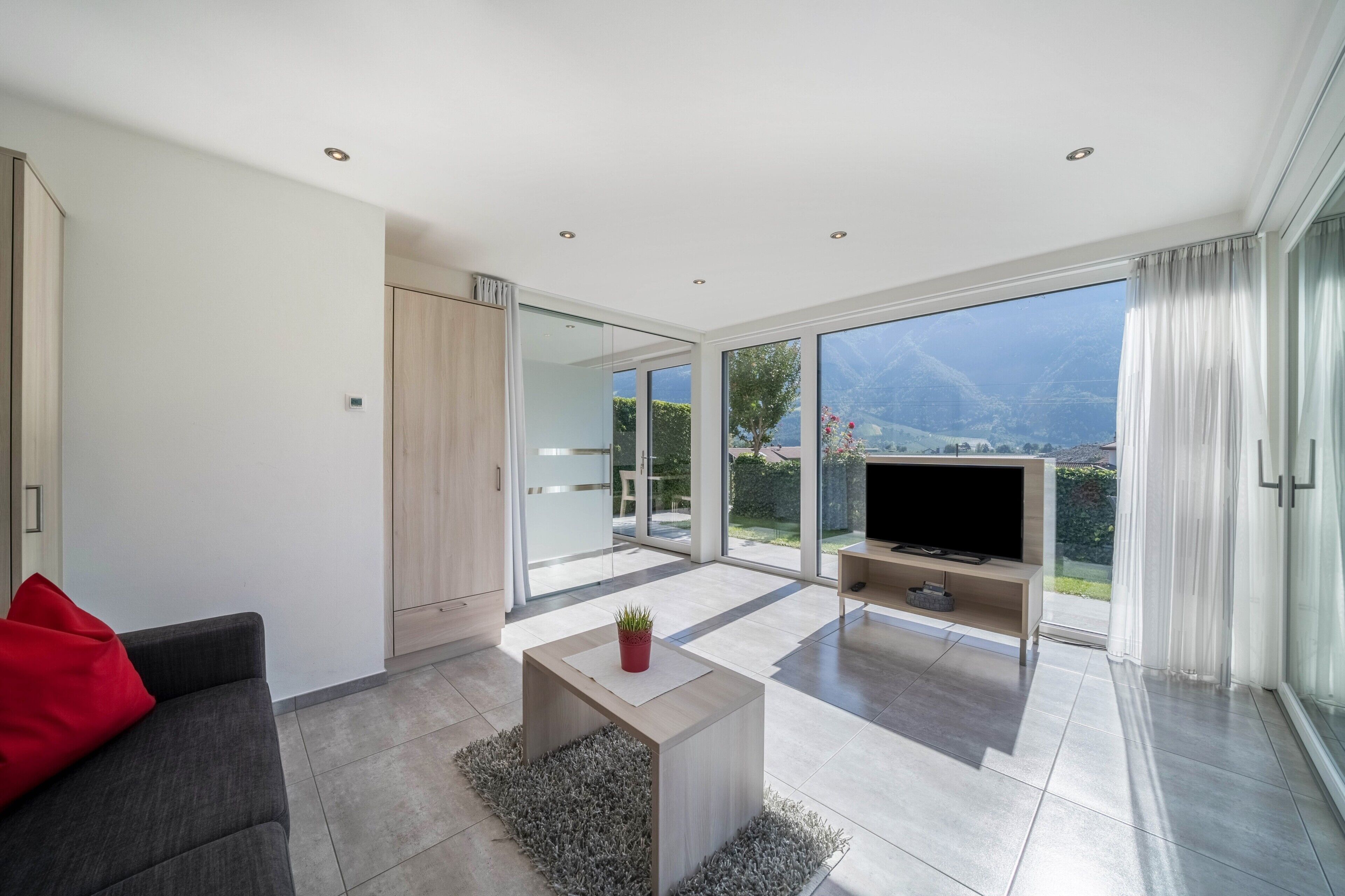 Ein modernes Wohnzimmer mit einer Couch, einem Couchtisch, einem Fernseher und einer Schiebetür zu einem Balkon mit Bergblick.