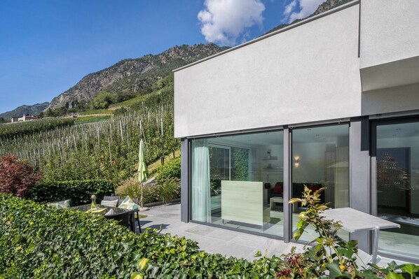 Un bâtiment moderne doté de grandes fenêtres en verre, d’un patio aménagé avec du mobilier d’extérieur et surplombant un paysage montagneux.