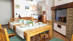 Zona pranzo con tavolo in legno apparecchiata per quattro persone, circondata da sedie. Sullo sfondo si intravede una cucina con mobili e fornello in legno.