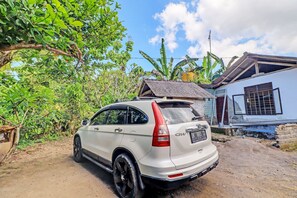A white SUV parked on a dirt road with a house and banana trees in the background. - Hotel O Pondok Ai (Negara)