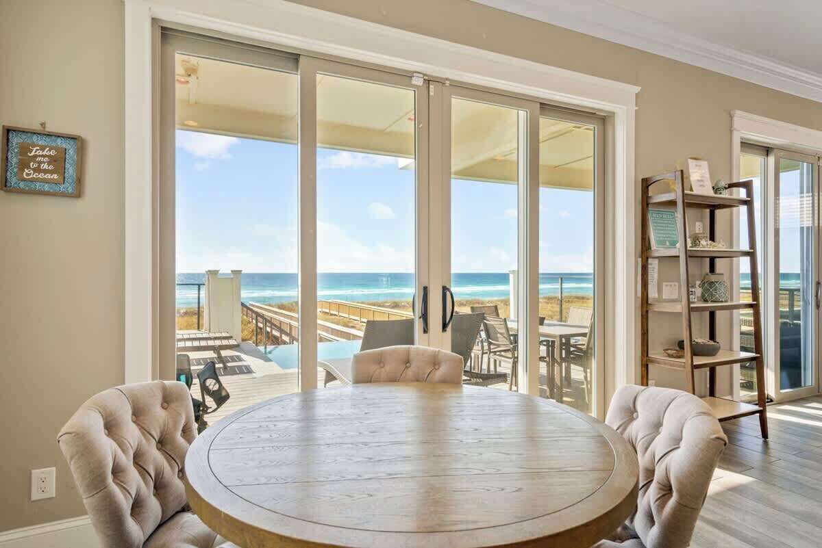 A round wooden table with two upholstered chairs, a sliding glass door leading to a deck with a view of the beach and ocean, and a bookshelf with decorative items.
