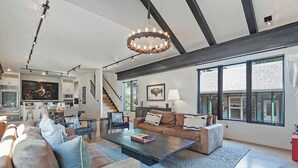 Ein geräumiges Wohnzimmer mit einer großen Couch, einem Couchtisch und einem Kamin. Eine Treppe führt zu einer oberen Ebene, und im Hintergrund ist eine Küche zu sehen.