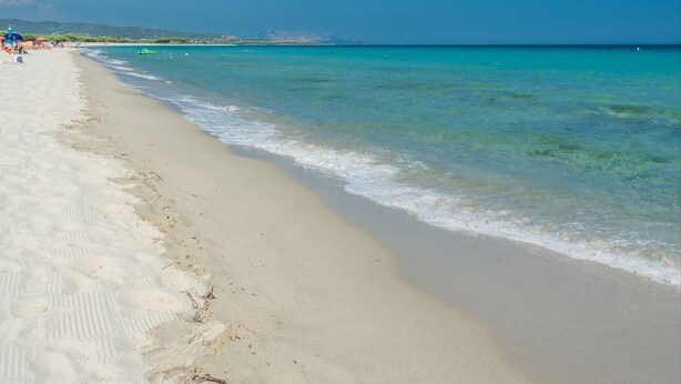 Une plage à l’eau turquoise et limpide, avec un rivage sablonneux et des collines qui se dessinent à l’horizon.