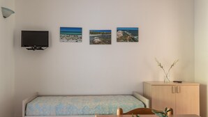 Une petite pièce comprenant un lit, un téléviseur fixé au mur et trois tableaux représentant des paysages côtiers.