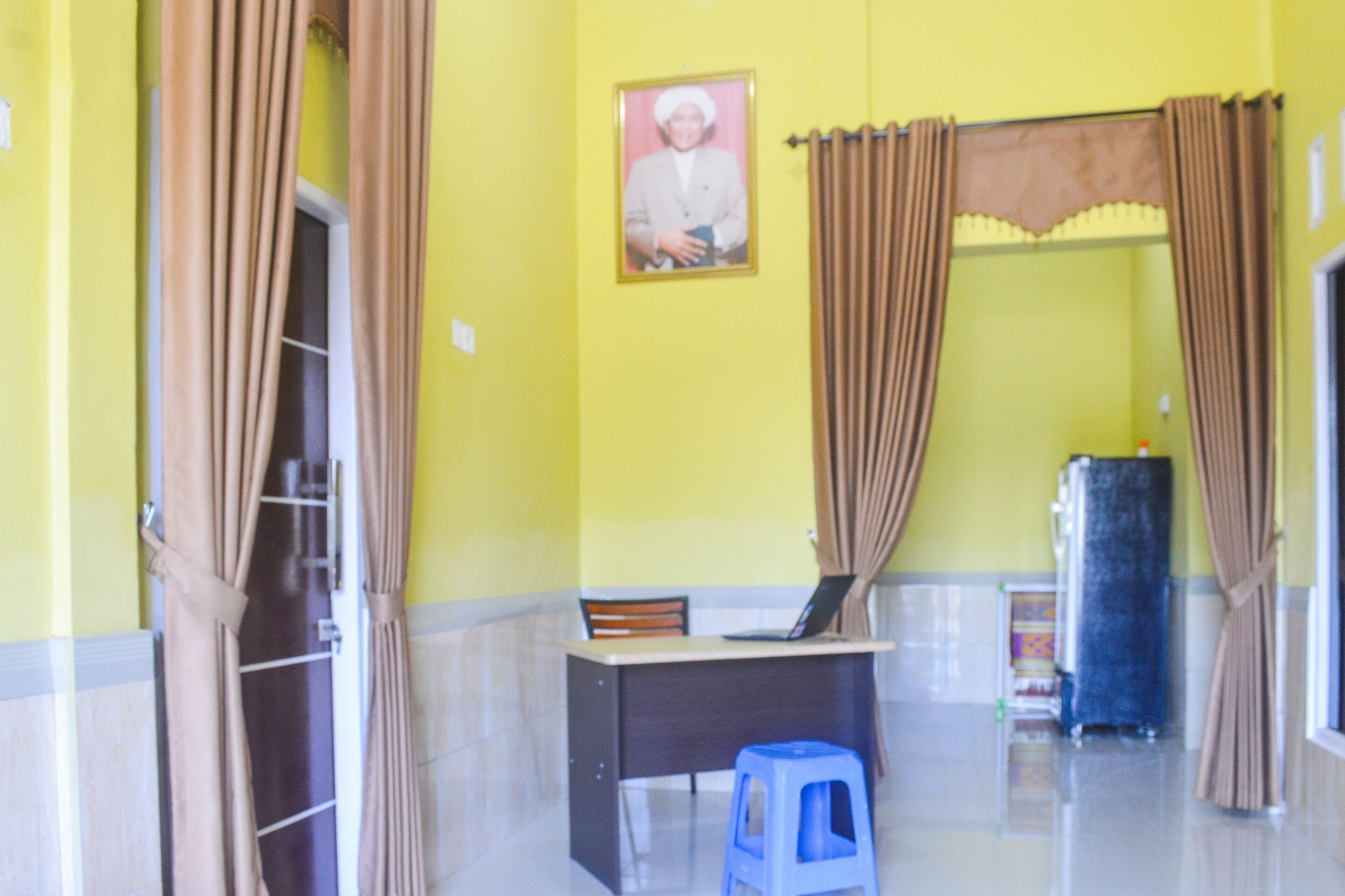 A room with yellow walls, a desk with a laptop, a blue stool, and a refrigerator.