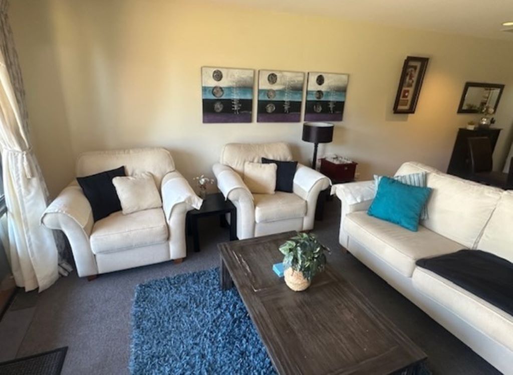 Een woonkamer met twee beige banken, een salontafel en een blauw vloerkleed. Er hangen drie schilderijen aan de muur en er staat een vloerlamp.