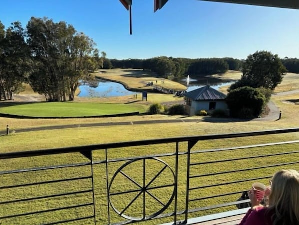 Vista desde un balcón sobre un campo de golf con un estanque, árboles y un pequeño edificio.