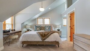 A bedroom with a sloped ceiling, a bed with a white comforter and patterned pillows, a wooden dresser, a desk with a lamp, and a window with blinds.