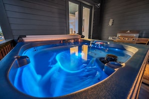 Ein Whirlpool mit blauer Beleuchtung, ein Grill und im Hintergrund der Blick auf das Haus.