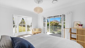 A bedroom with a large bed, a wicker chair, and a view of a garden through open windows. - Hygge House - Kangaroo valley (Kangaroo Valley)