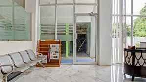 Um hall com balcão de recepção, área de espera com cadeiras e porta de vidro que dá para uma área externa.