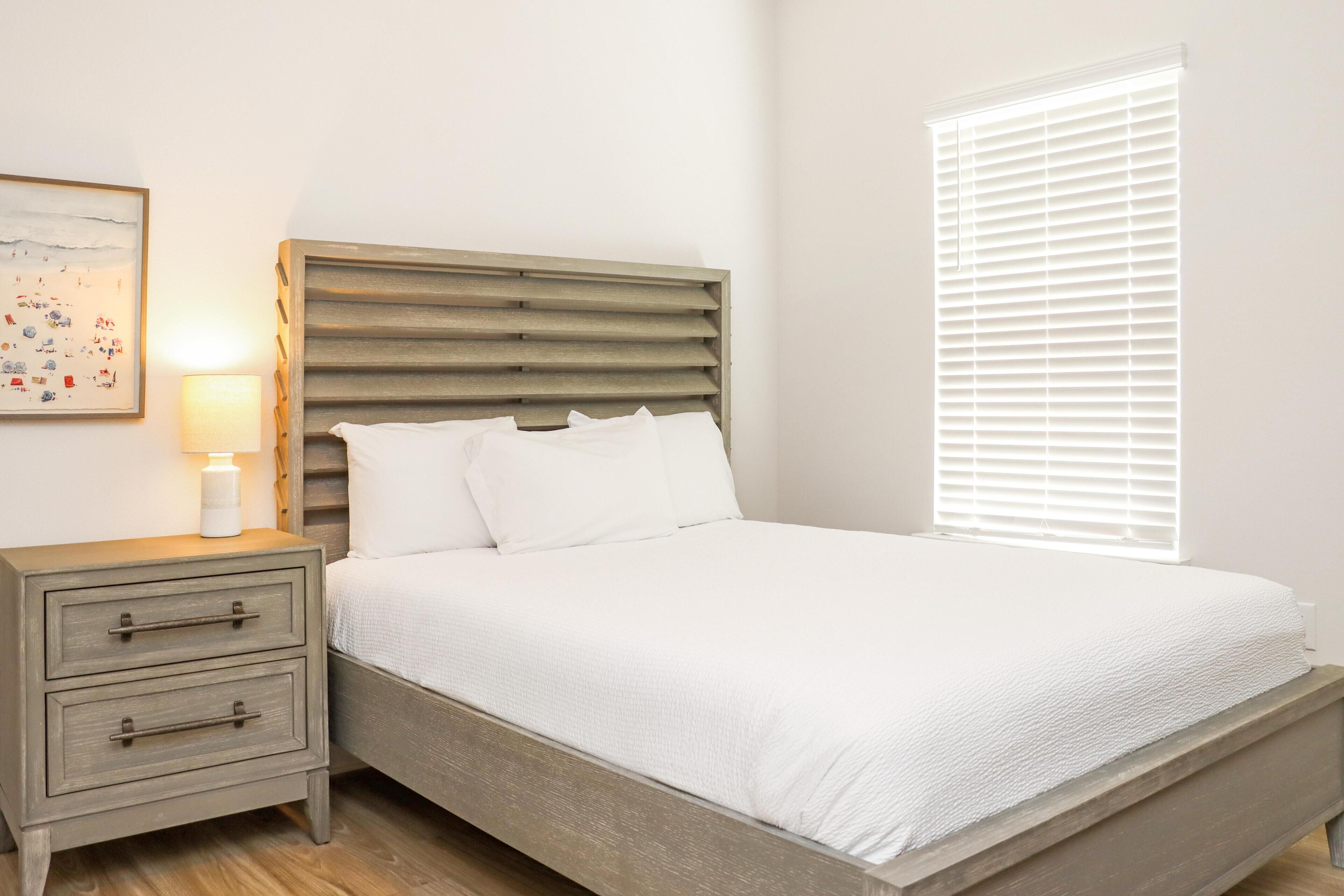 Quarto com cama de madeira, roupa de cama branca, mesa de cabeceira com abajur e janela com persianas.