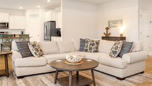 A living room with a beige sectional sofa, a round coffee table, and a kitchen in the background. - Beach Haven at WindMark (Port St. Joe)