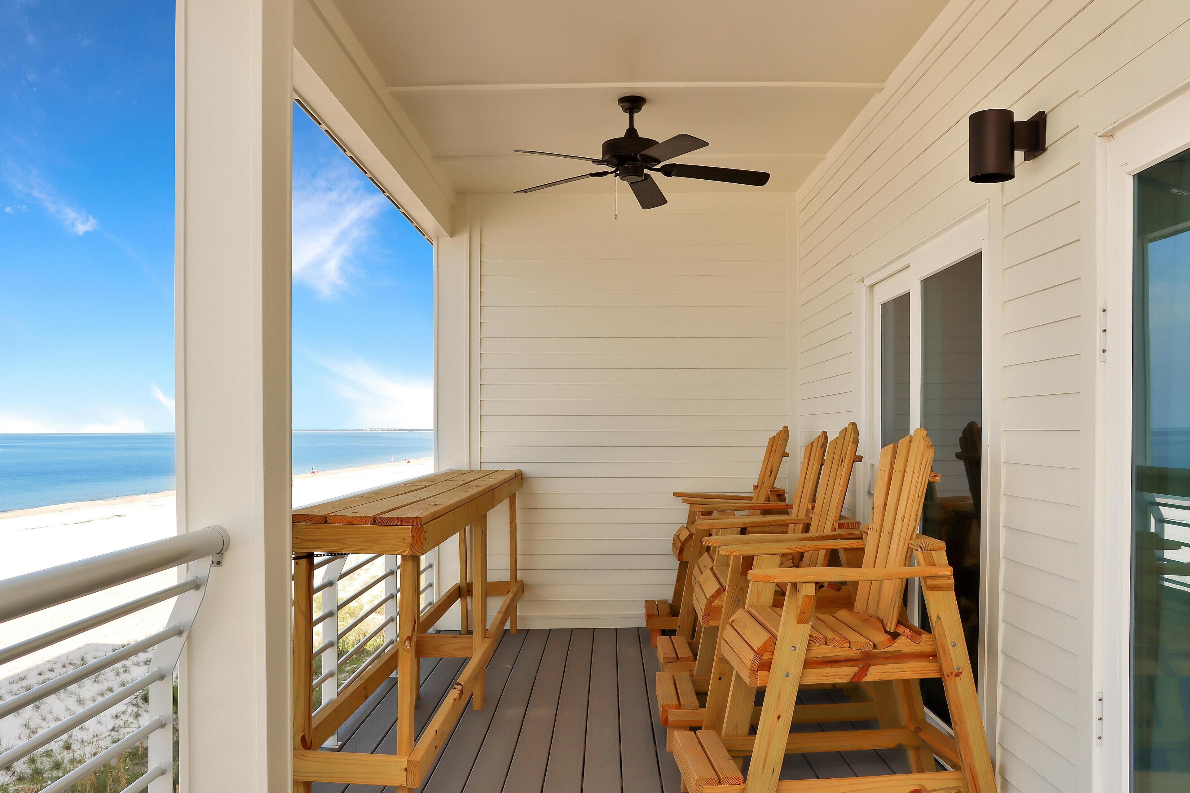 Un balcone con ventilatore a soffitto, sedie in legno e un tavolo vista spiaggia.