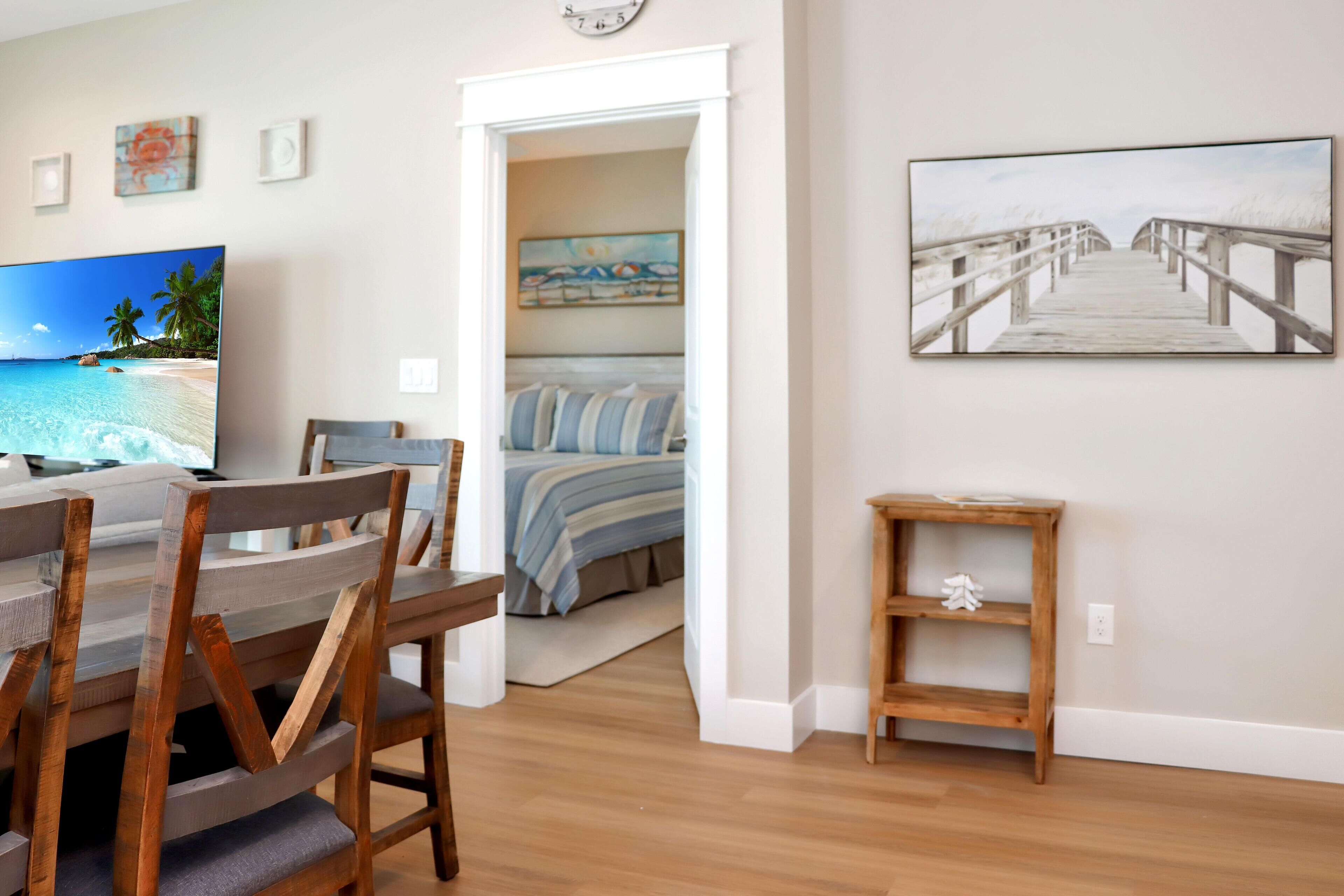 A room with a wooden dining table, chairs, a bed with striped bedding, a painting of a beach, and a wooden bench.