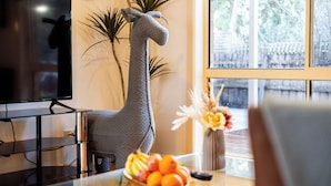 A living room with a large giraffe sculpture, a television, a dining table with fruit, and a window with a view of trees. - New Melbourne Staycation 3BRM (Glen Waverley)