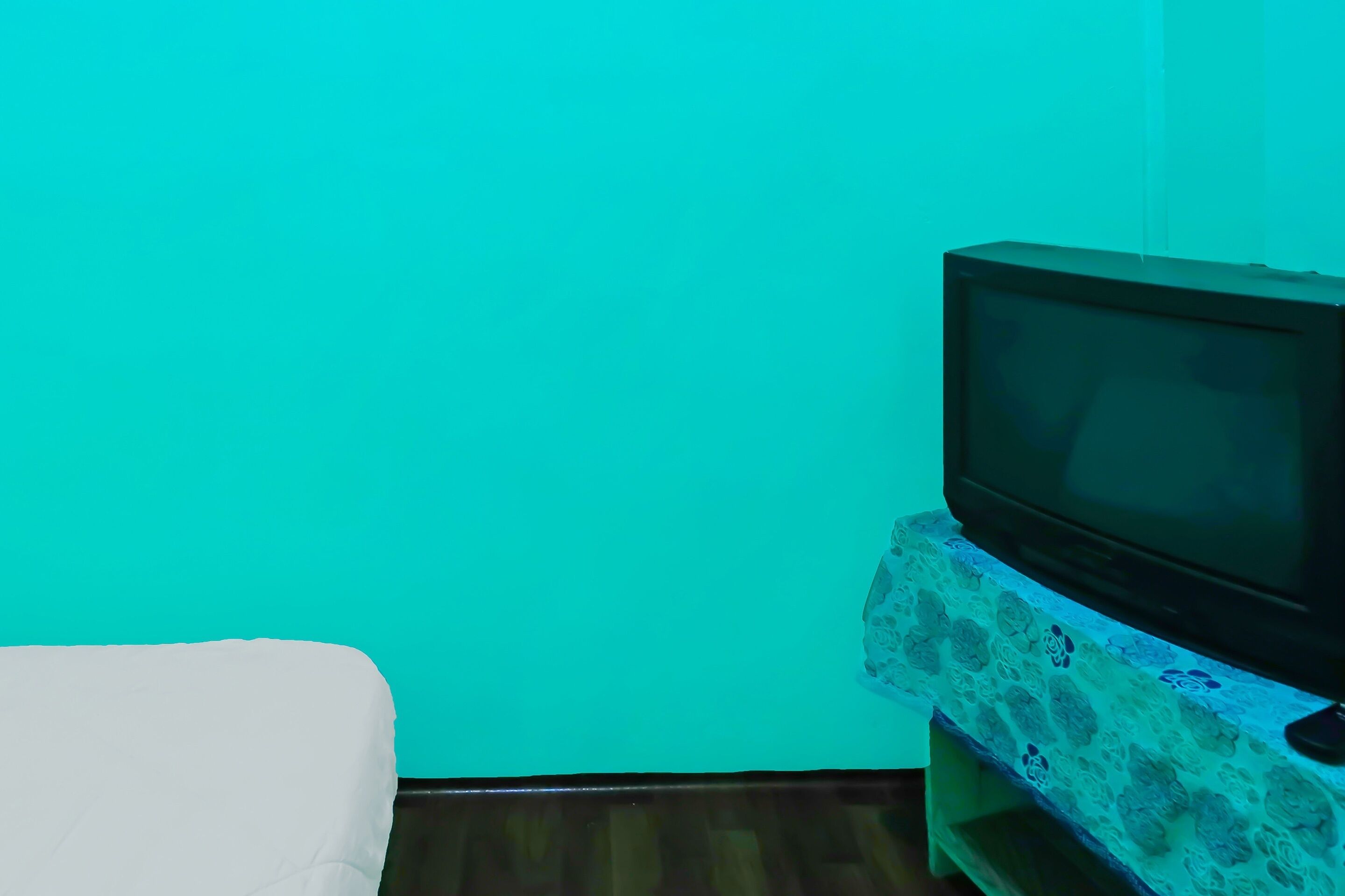 Una habitación con pared verde azulado, un televisor sobre un mueble estampado con motivos florales y una cama blanca.